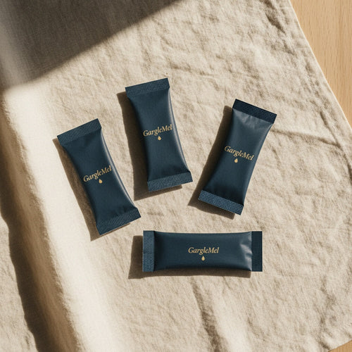 Four GargleMel sachets — navy paper stick-packs with gold wordmark — on cream linen